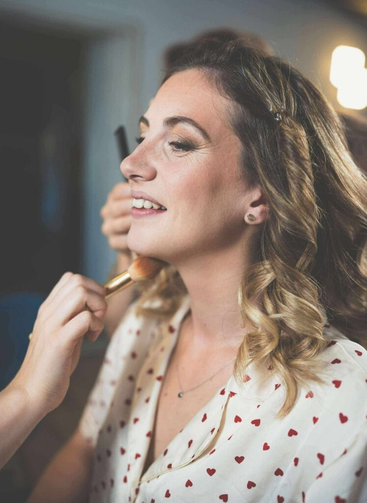 Femme souriante en préparation mariage recevant un maquillage naturel avec pinceau teint et coiffure ondulée.