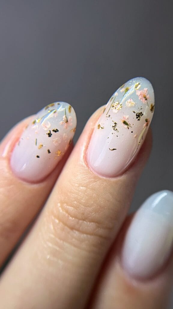 Manucure nude rosée avec petites fleurs séchées et feuilles d’or sur ongles amande brillants.