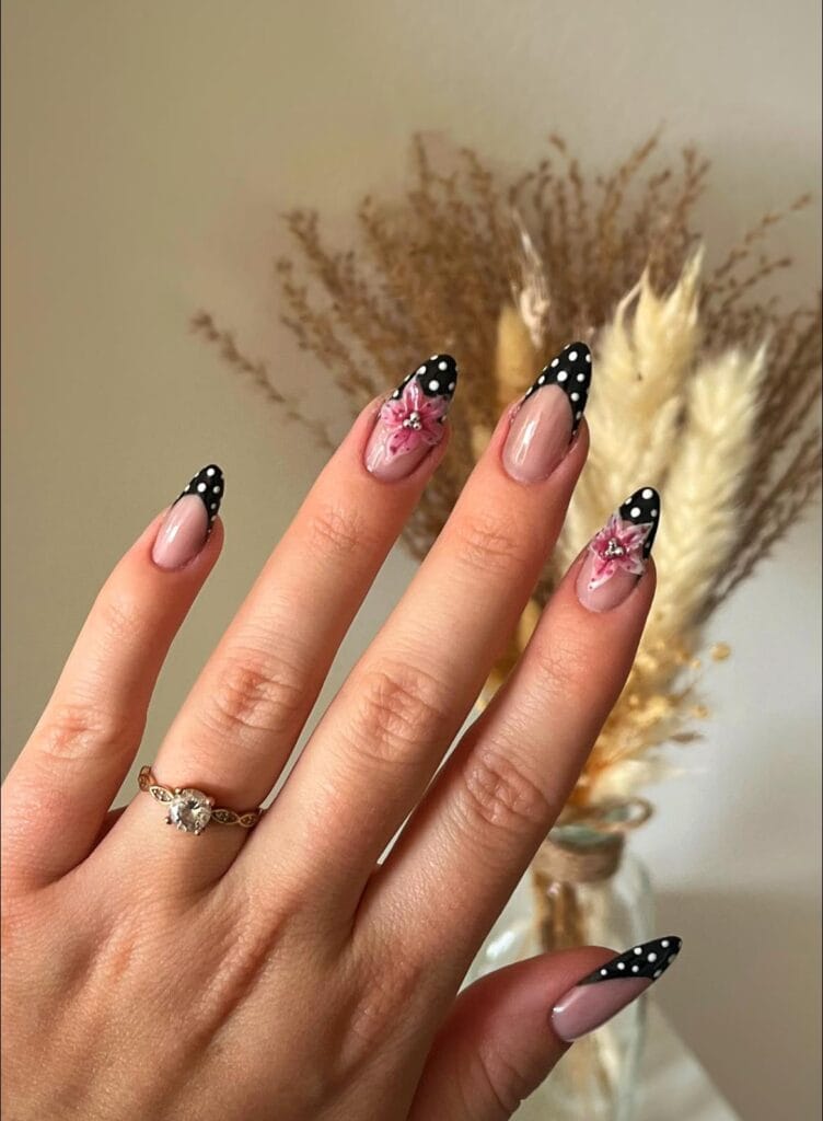 Pose d’ongles amande avec french noire à pois blancs et fleurs roses 3D sur base nude brillante.